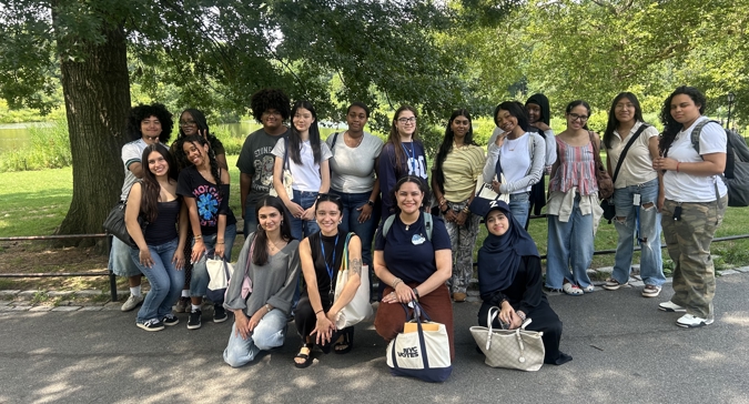 Group photo of the 2026 Youth Ambassadors, about 18 teens and young adults, posed together on a paved park path under large shade trees, with a pond and greenery in the background. Several people hold NYC Votes tote bags.