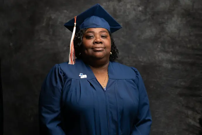 A CCF graduate wearing a navy blue cap and gown stands in front of a dark textured backdrop, with a CCF pin on the front of the gown.​​ 