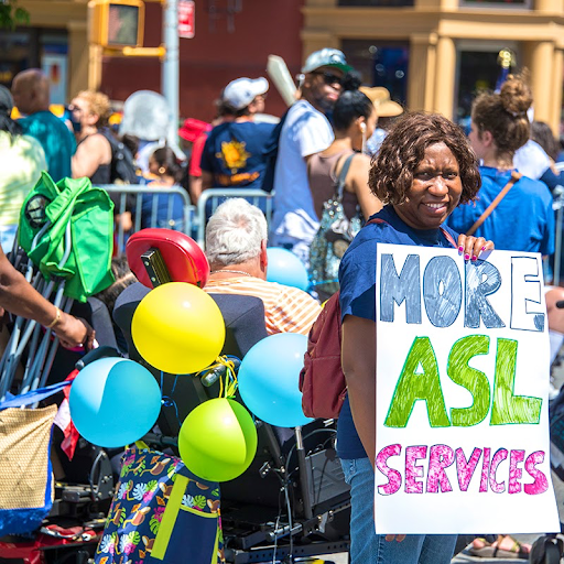 Un participant à la Parade de la fierté des personnes en situation de handicap de 2024 tient une pancarte sur laquelle on peut lire : « Plus de services ASL » .