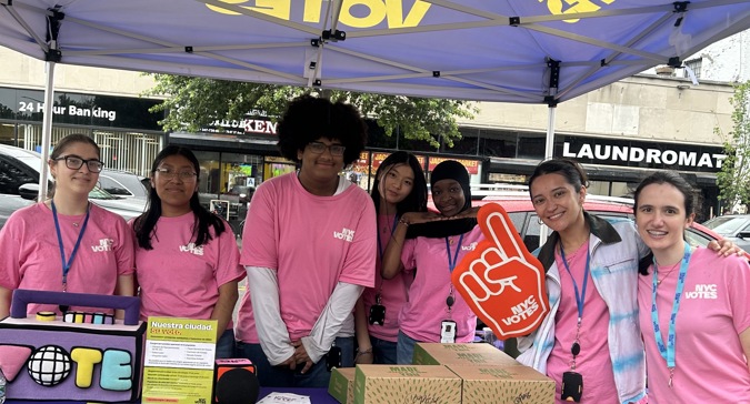 María holds an NYC Votes foam finger while posing with Youth Ambassadors at an NYC Votes outreach table during Primary Election Day in Jackson Heights. The group wears NYC Votes shirts and stands beneath an NYC Votes tent, with voter registration materials and outreach supplies displayed on the table.