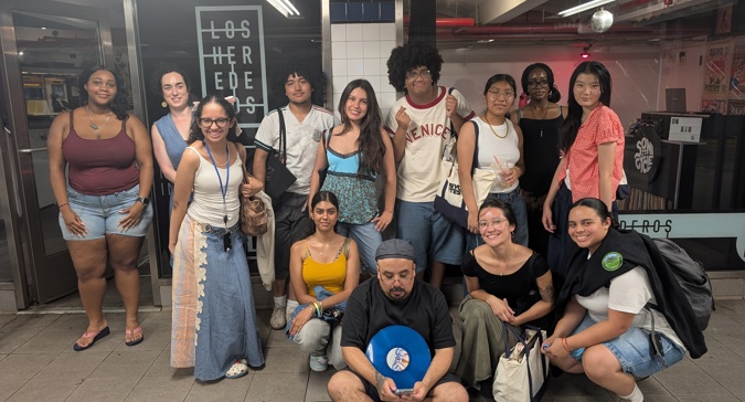 María, NYC Votes Youth Program Coordinator, poses with the 2026 Youth Ambassadors at Los Herederos Radio Station inside the 74th Street–Jackson Heights subway station. The group of 12 stands and sits together, smiling at the camera, with one person holding a blue record and another carrying an NYC Votes tote bag.