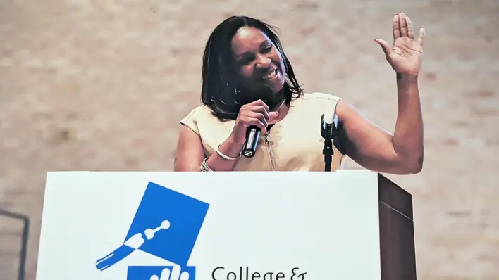 A speaker stands at a podium displaying the College & Community Fellowship logo, holding a microphone with one hand raised.​​ 