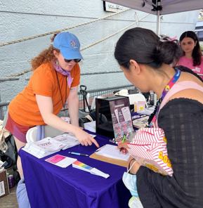 Two CFB staff stand smiling behind a podium at an outdoor community event under a blue canopy tent. A sign on the podium reads “Latino Week of Action | La Voz de Mi Gente.” One woman wears a burgundy dress and gray cardigan, while the other wears an orange “NYC Votes Staff” T-shirt. Storefronts, additional tents, and a city sidewalk are visible in the background.