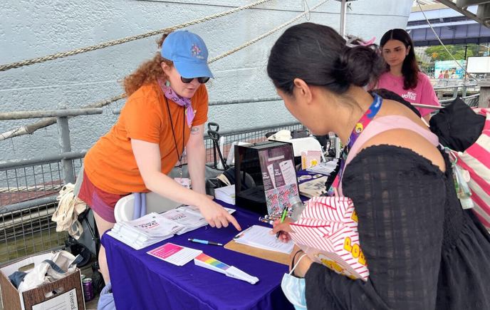 Two CFB staff stand smiling behind a podium at an outdoor community event under a blue canopy tent. A sign on the podium reads “Latino Week of Action | La Voz de Mi Gente.” One woman wears a burgundy dress and gray cardigan, while the other wears an orange “NYC Votes Staff” T-shirt. Storefronts, additional tents, and a city sidewalk are visible in the background.
