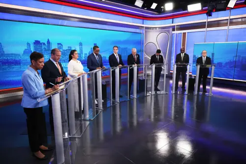 Nine candidates in front of podiums on a stage with a blue backdrop of the city skyline