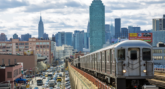 Elevated subway train traveling above a busy street in Queens, with traffic below and the Empire State Building visible in the New York City skyline in the background.​​ 
