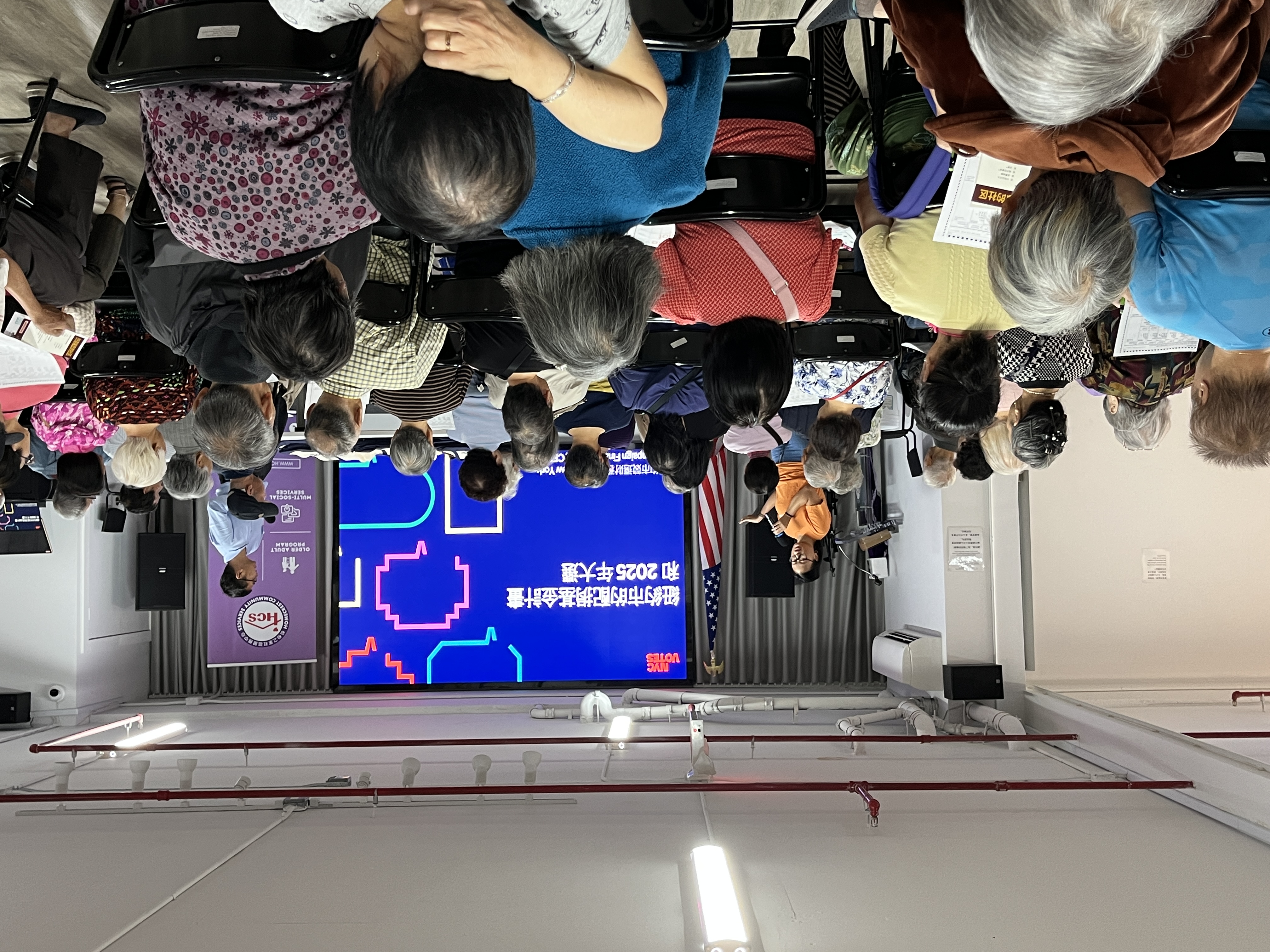 A presenter speaks into a microphone at the front of a crowded community room, addressing an audience of mostly older adults seated in rows. A large screen behind them displays a presentation in Chinese about NYC Votes and the 2025 election, with colorful graphics. An American flag stands to the side, and another facilitator stands near the screen.​​  