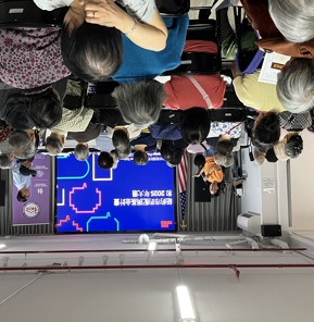 A presenter speaks into a microphone at the front of a crowded community room, addressing an audience of mostly older adults seated in rows. A large screen behind them displays a presentation in Chinese about NYC Votes and the 2025 election, with colorful graphics. An American flag stands to the side, and another facilitator stands near the screen.​​ 