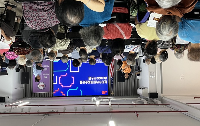 A presenter speaks into a microphone at the front of a crowded community room, addressing an audience of mostly older adults seated in rows. A large screen behind them displays a presentation in Chinese about NYC Votes and the 2025 election, with colorful graphics. An American flag stands to the side, and another facilitator stands near the screen.​​ 