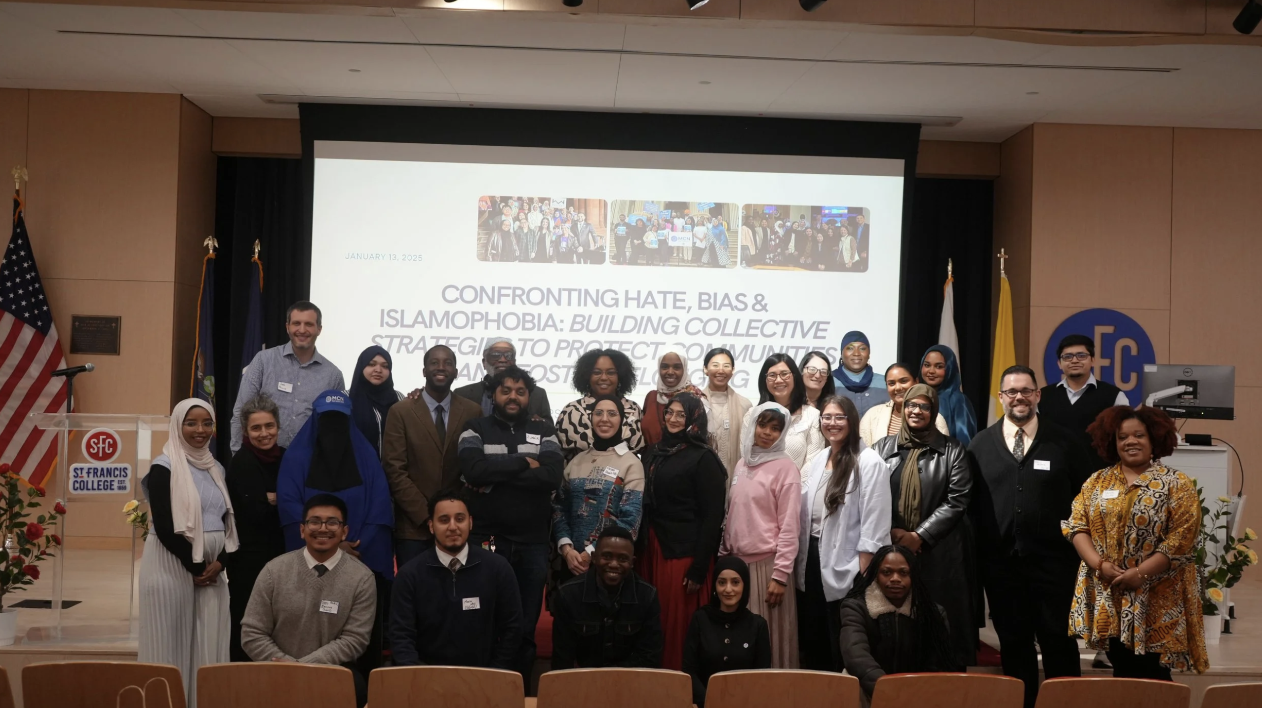 Large group of people posing for a photo in an auditorium at St. Francis College. They stand and kneel in several rows in front of a projection screen that reads, “Confronting Hate, Bias & Islamophobia: Building Collective Strategies to Protect Communities,” dated January 13, 2025. U.S. and New York State flags flank the stage, and an SFC logo is visible on the wall. Many attendees wear name tags and a mix of business, casual, and modest attire.​​ 