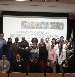 Large group of people posing for a photo in an auditorium at St. Francis College. They stand and kneel in several rows in front of a projection screen that reads, “Confronting Hate, Bias & Islamophobia: Building Collective Strategies to Protect Communities,” dated January 13, 2025. U.S. and New York State flags flank the stage, and an SFC logo is visible on the wall. Many attendees wear name tags and a mix of business, casual, and modest attire.​​ 