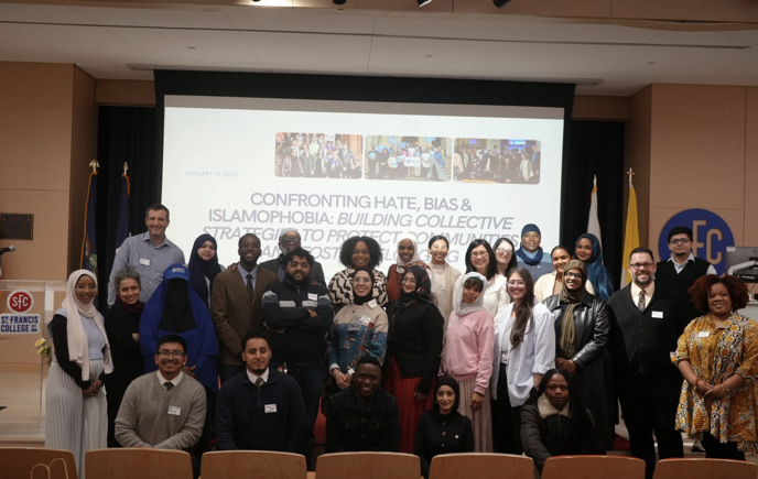 Large group of people posing for a photo in an auditorium at St. Francis College. They stand and kneel in several rows in front of a projection screen that reads, “Confronting Hate, Bias & Islamophobia: Building Collective Strategies to Protect Communities,” dated January 13, 2025. U.S. and New York State flags flank the stage, and an SFC logo is visible on the wall. Many attendees wear name tags and a mix of business, casual, and modest attire.