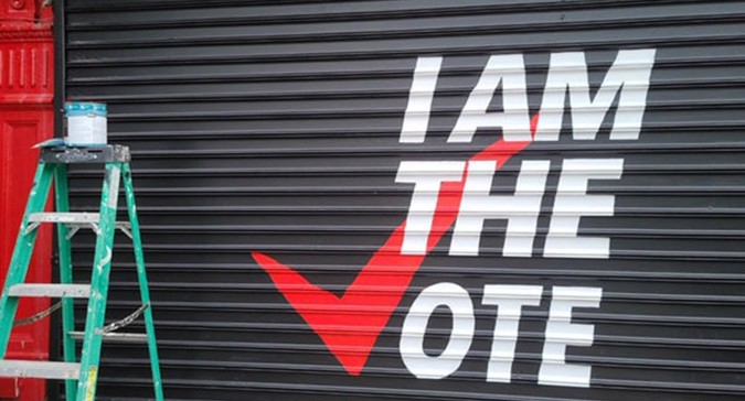 A business' garage door is pulled down revealing a mural that says "I am the Vote". the 'V' in 'Vote' is a red checkmark signifying a ballot checkmark. There is a green ladder with a bucket  of paint resting on top to the left of the garage door.