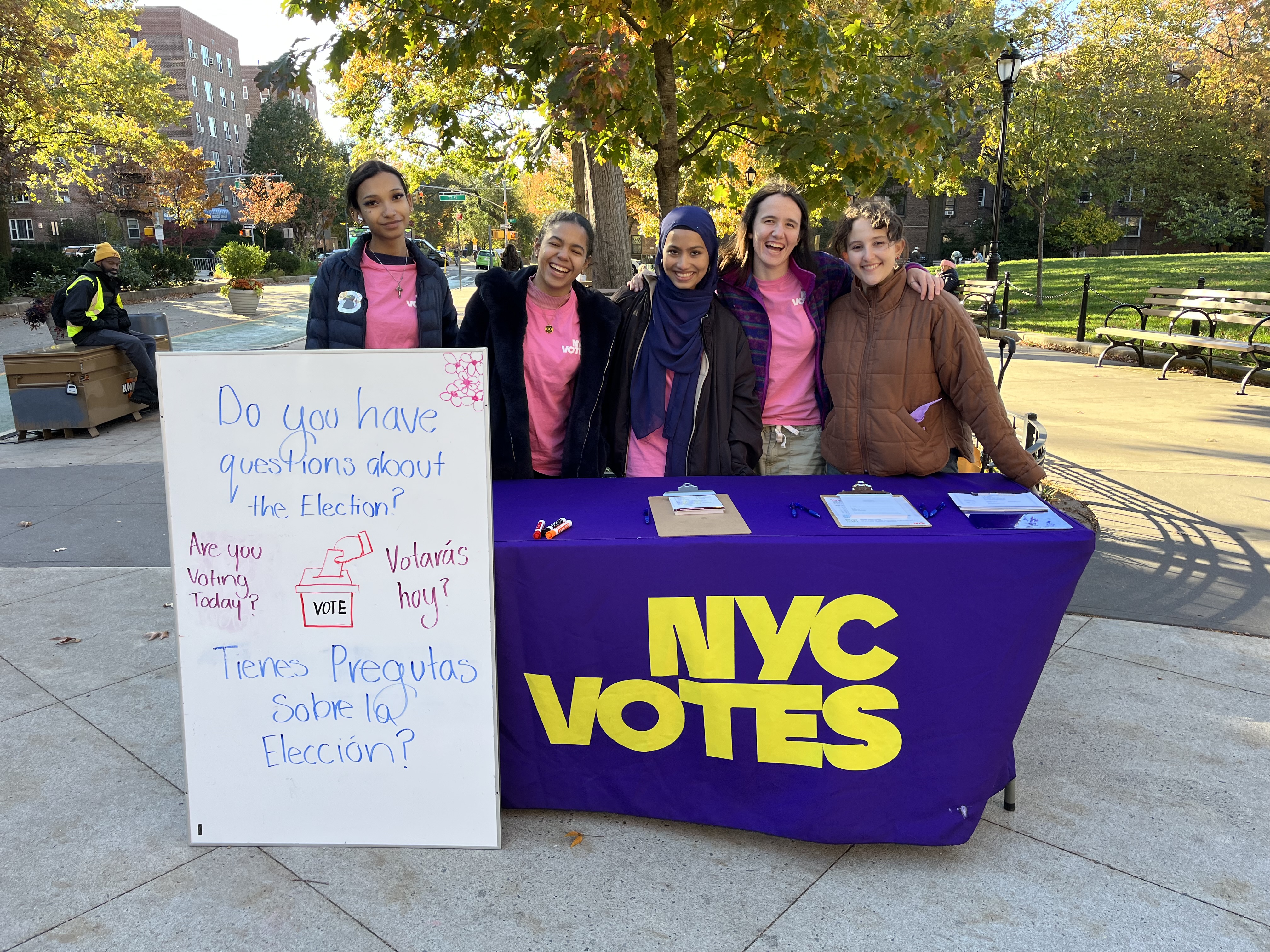 Five people standing behind an NYC Votes table outdoors in a park. They are smiling and posing for the camera. The table is covered with a purple cloth featuring the NYC Votes logo in large yellow letters. To the left of the table is a white sign with text in English and Spanish that reads "Do you have questions about the Election? Are you Voting Today? Tienes Preguntas Sobre la Elección? Votará hoy?" The background shows trees with autumn leaves, park benches, and city buildings.​​ 