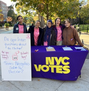 Five people standing behind an NYC Votes table outdoors in a park. They are smiling and posing for the camera. The table is covered with a purple cloth featuring the NYC Votes logo in large yellow letters. To the left of the table is a white sign with text in English and Spanish that reads "Do you have questions about the Election? Are you Voting Today? Tienes Preguntas Sobre la Elección? Votará hoy?" The background shows trees with autumn leaves, park benches, and city buildings.​​ 
