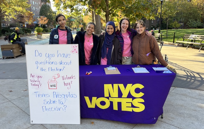Five people standing behind an NYC Votes table outdoors in a park. They are smiling and posing for the camera. The table is covered with a purple cloth featuring the NYC Votes logo in large yellow letters. To the left of the table is a white sign with text in English and Spanish that reads "Do you have questions about the Election? Are you Voting Today? Tienes Preguntas Sobre la Elección? Votará hoy?" The background shows trees with autumn leaves, park benches, and city buildings.​​ 