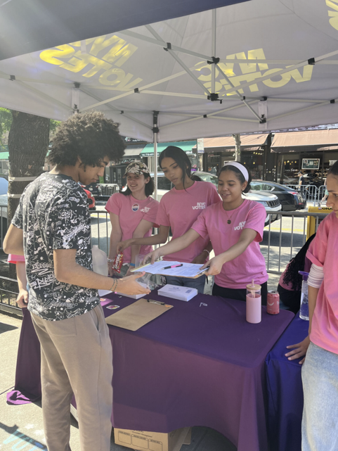 A group of young NYC Votes ambassadors in pink t-shirts working under a white branded tent. One ambassador is handing a clipboard to a young man in a patterned shirt to help him register or find election information.​​ 