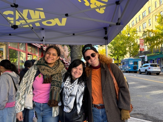 Three CFB staff members stand together under an NYC Votes canopy tent at an outdoor street event. They are wearing layered clothing suitable for cool weather, including jackets, scarves, and a knit cap. Behind them, other NYC Votes staff are gathered near storefronts. The street next to them has parked cars, a blue food truck, and buildings lined with trees showing green foliage.