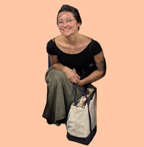 María, NYC Votes Youth Programs Coordinator, smiling while crouched, wearing a black top and olive pants, with a canvas tote bag beside her.