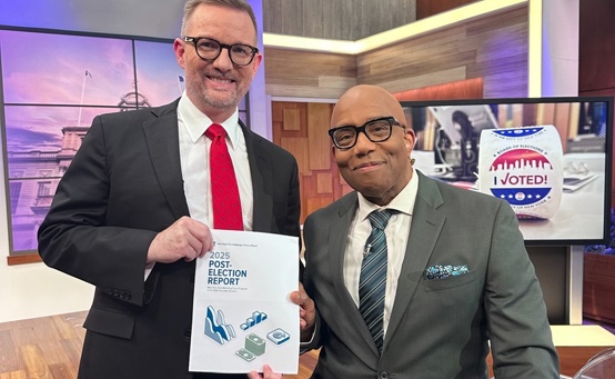 The executive director of C F B stands next to a seated journalist while the executive director holds a copy of the 2025 post-election report