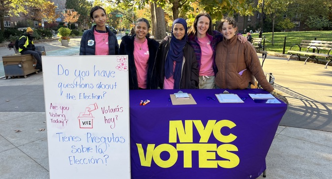 A group of volunteers in pink “NYC Votes” shirts hand paperwork to a person at an outdoor voter registration table on a city sidewalk.